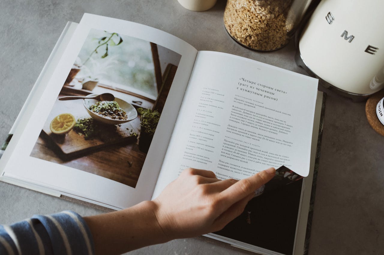 services-01 A person turns the page of a recipe book on a kitchen counter, surrounded by cooking ingredients.